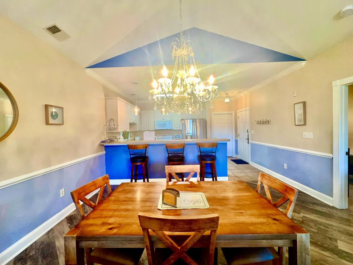 An inviting dining area featuring a large dining table under an elegant chandelier, with a view of the kitchen bar area in the background. The setting is perfect for family meals, celebrations, and shared experiences.