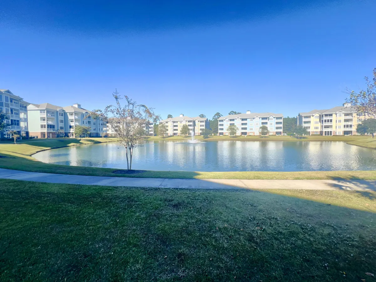A tranquil pond bordered by landscaped lawns, with residential buildings and mature trees creating a peaceful, inviting setting. The image captures the open space where families can enjoy an afternoon stroll or gather for outdoor relaxation.