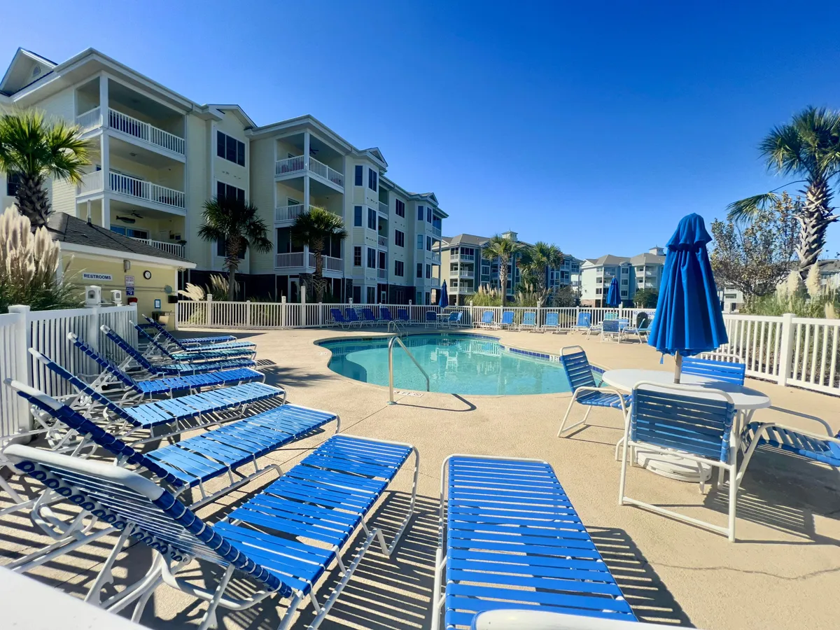 A sparkling blue swimming pool surrounded by sun loungers, umbrellas, and palm trees, creating a cheerful and relaxing resort ambiance. The area is ideal for both swimming and lounging, with plenty of space for families.