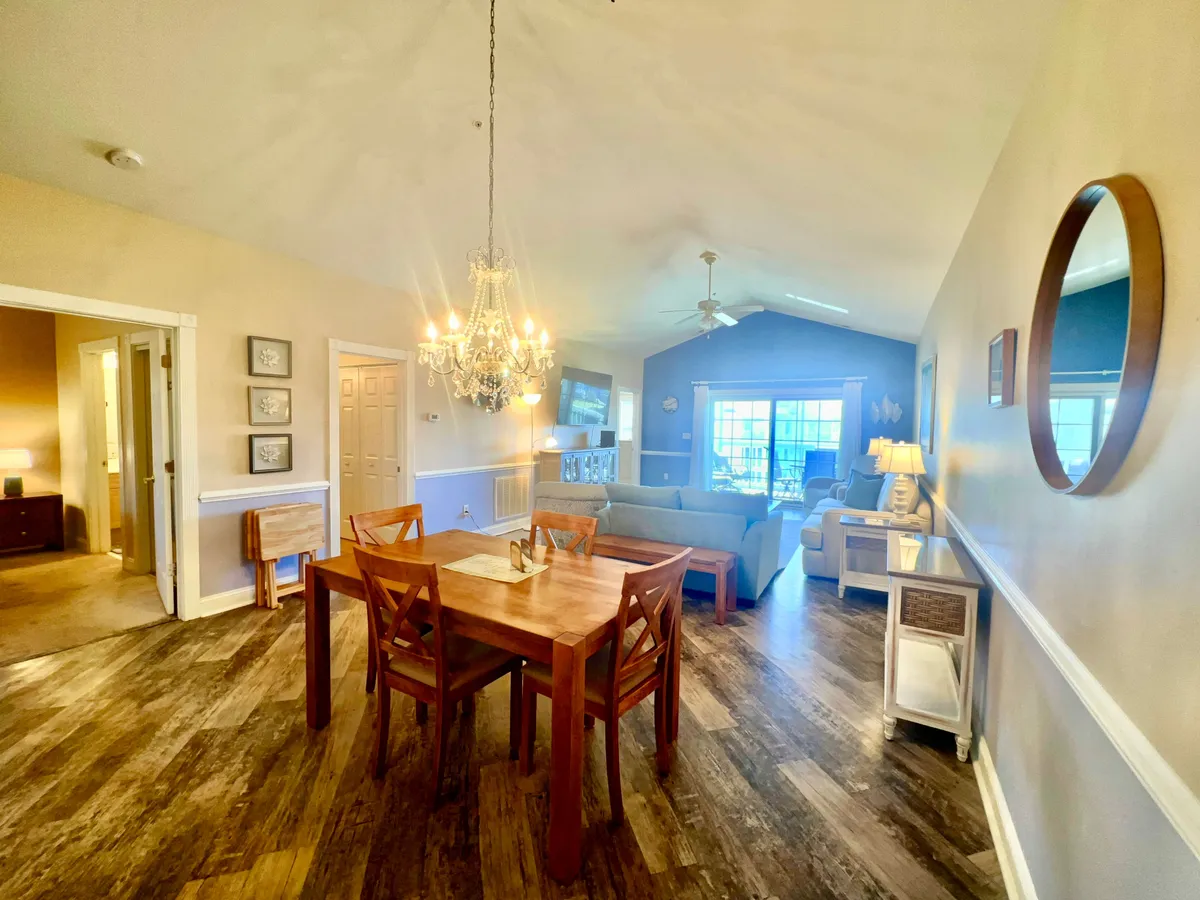 This image captures the open-concept living and dining space, prominently featuring a plush sofa, an elegant dining table under a chandelier, and expansive windows that allow natural light to flood the area. The windows provide tranquil views and the setup effortlessly blends comfort with welcoming communal spaces.