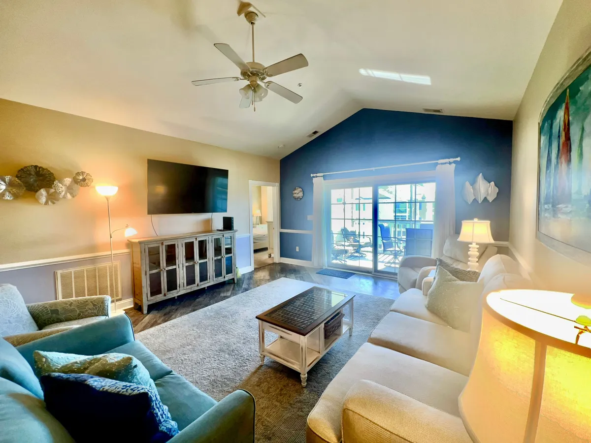 The scene features a comfortable living area with a plush sofa set, a coffee table, a television, and large windows that open to outdoor views. The setup suggests a space ideal for family movie nights, relaxed conversations, or enjoying the scenery outside.