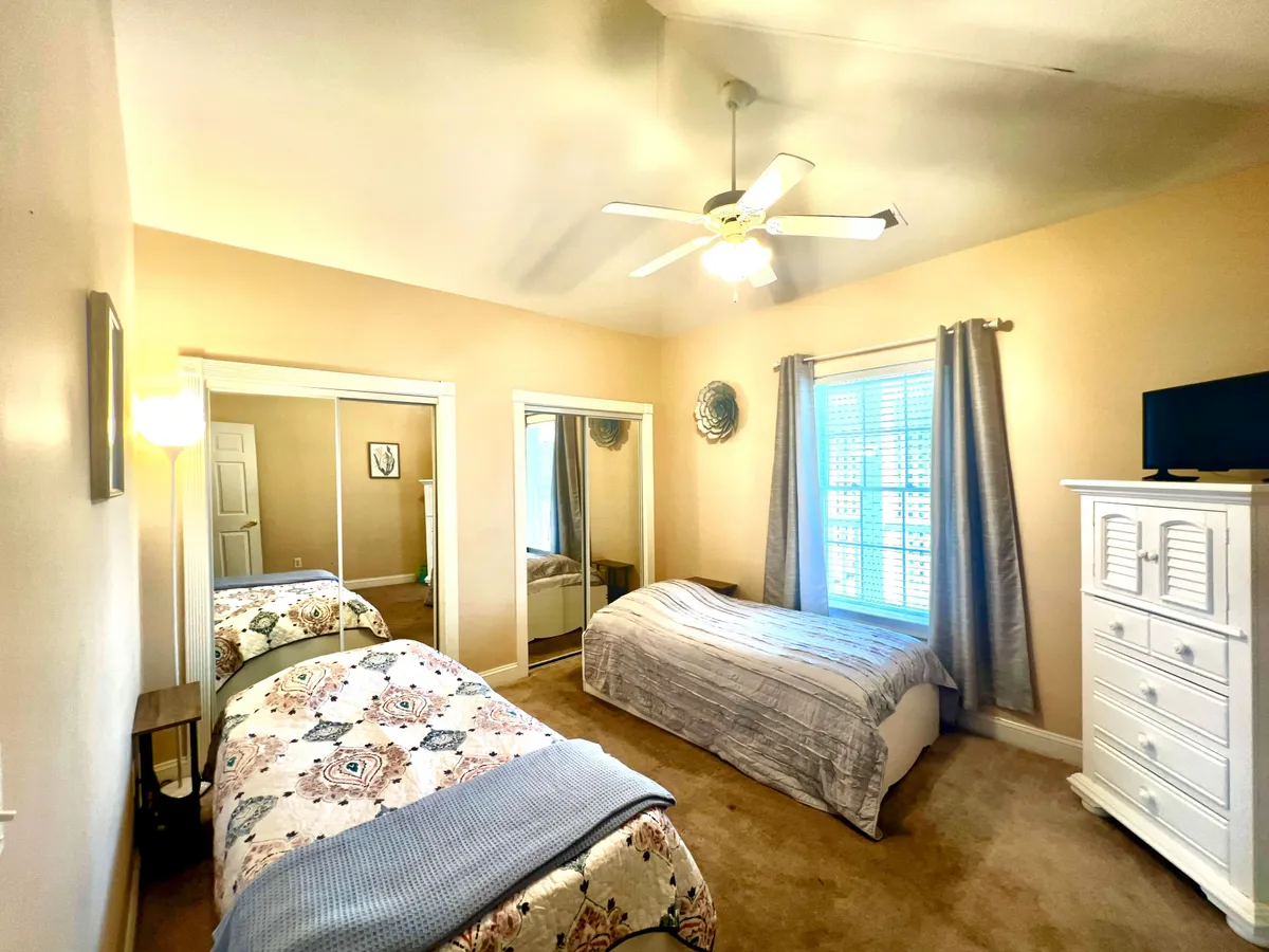 A spacious bedroom with two beds, a large mirror, a window with curtains, and a dresser topped with a TV. The room glows with morning sunlight, inviting guests to relax, read, or enjoy cozy evenings together.