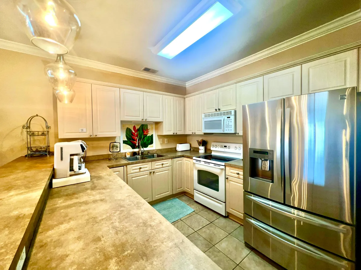 This image features a welcoming kitchen with a stainless steel refrigerator, modern appliances, a decorative plant, and stylish light fixtures. The countertops are spacious, and there is an overall sense of functionality and warmth, ideal for both casual and special meal prep.