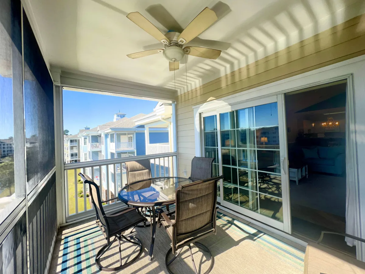 A cozy screened balcony with a glass table and four chairs invites families to gather for morning coffee or evening chats. The ceiling fan hints at comfort, while the open-air setting connects directly to the outdoors.