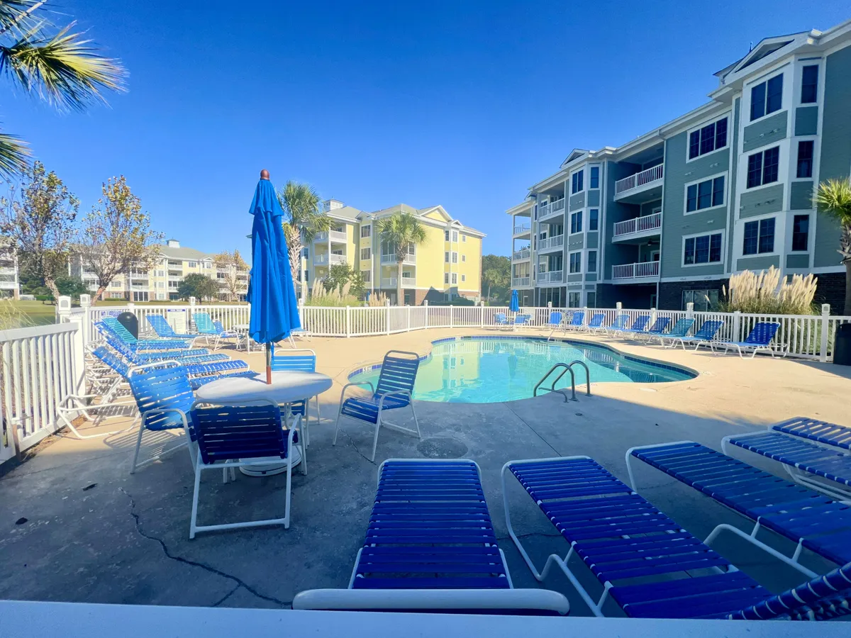 A resort-style swimming pool surrounded by lounge chairs and umbrellas, with nearby buildings providing a welcoming backdrop. The scene suggests leisure, socializing, and fun under the sun—perfect for family members of all ages.