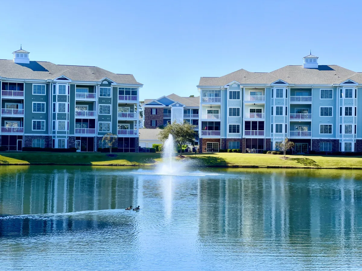 A tranquil lake with a picturesque fountain is bordered by apartment buildings, while ducks glide across the water. The landscaped grounds suggest a peaceful place for family walks or sunset strolls.