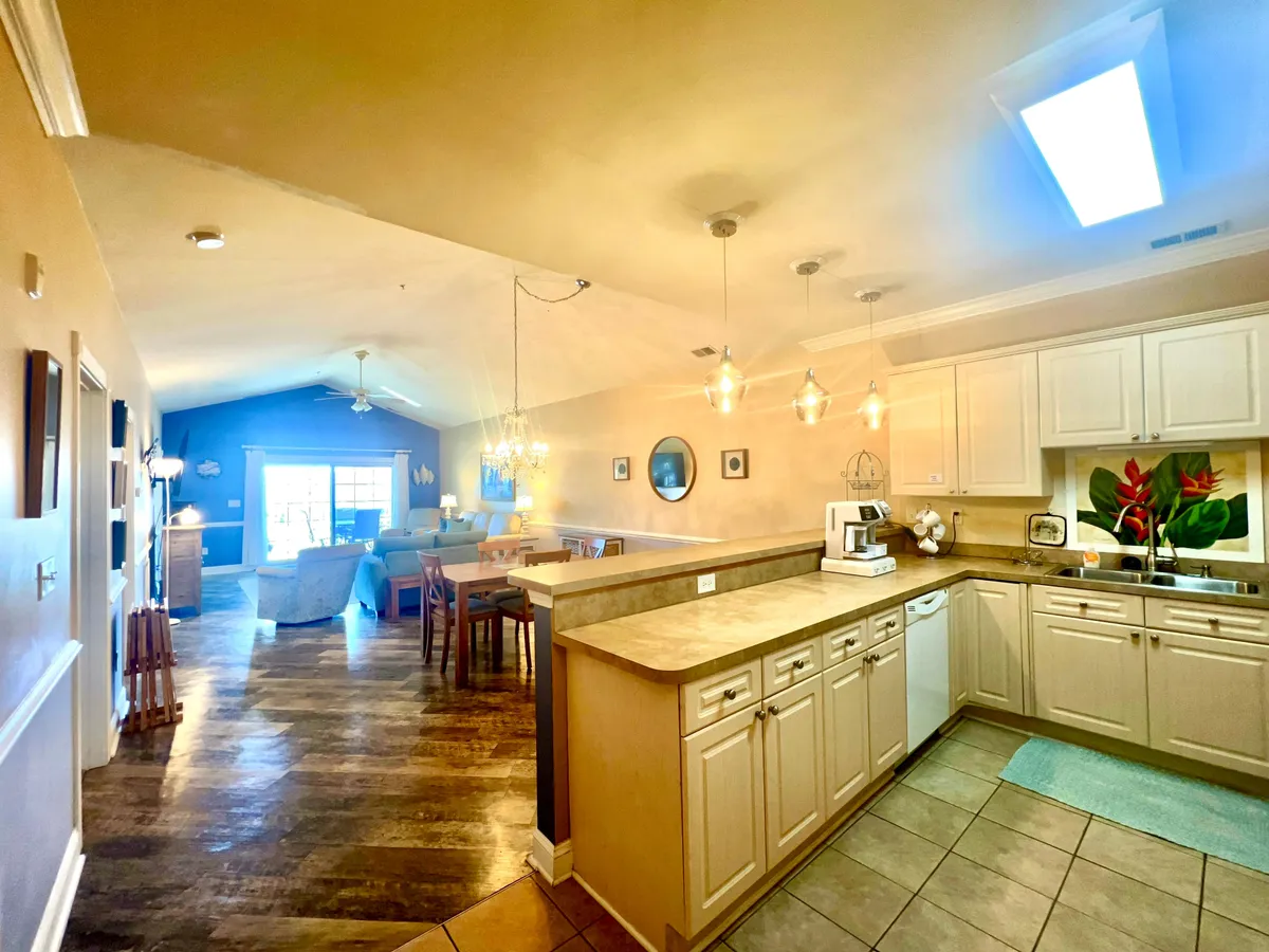 A wide view of an inviting, family-friendly living and dining space. The image includes a kitchen island, a cozy living room seating area, a spacious dining table, and a large window letting in abundant sunlight. Decorative plants add warmth and a homelike touch.