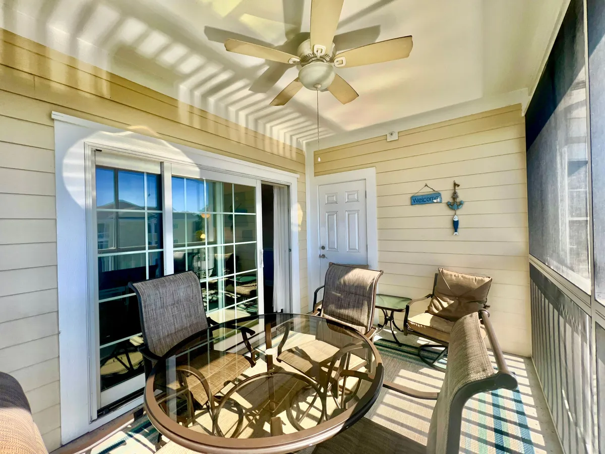 A round glass table and comfortable chairs are set in a well-lit area with a ceiling fan and a ‘welcome’ sign, suggesting a relaxed spot for casual dining, morning coffee, or evening gatherings. Natural light and an airy feel underscore its versatility.